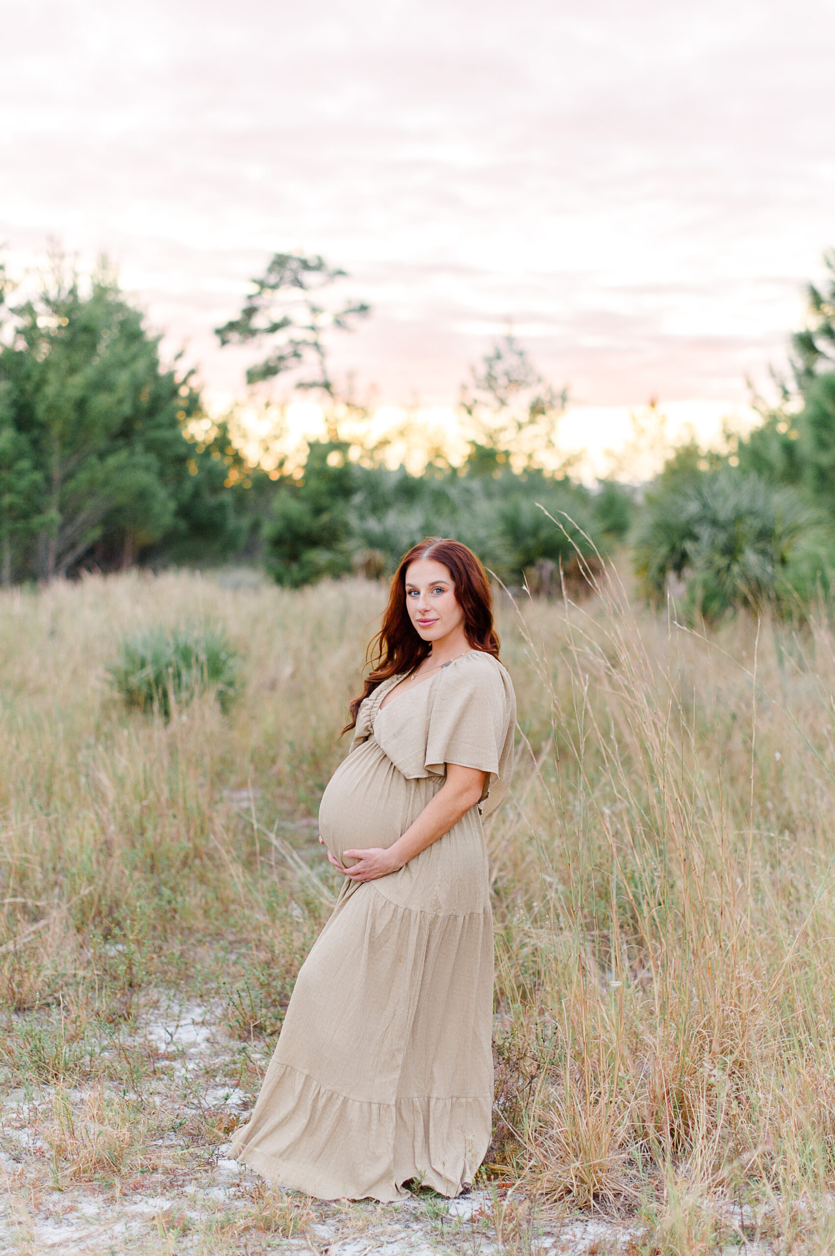 Pregnant mother smiles at the camera while holding her belly in a tall grass field during her Maternity photo session with M. Lauren Photography Orlando prenatal yoga