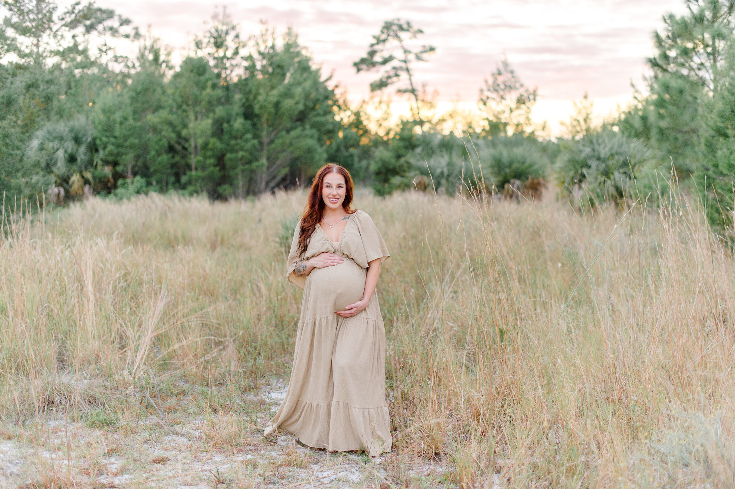 Pregnant mother stands in a field at sunset holding her belly and smiling at the camera Orlando Prenatal Yoga