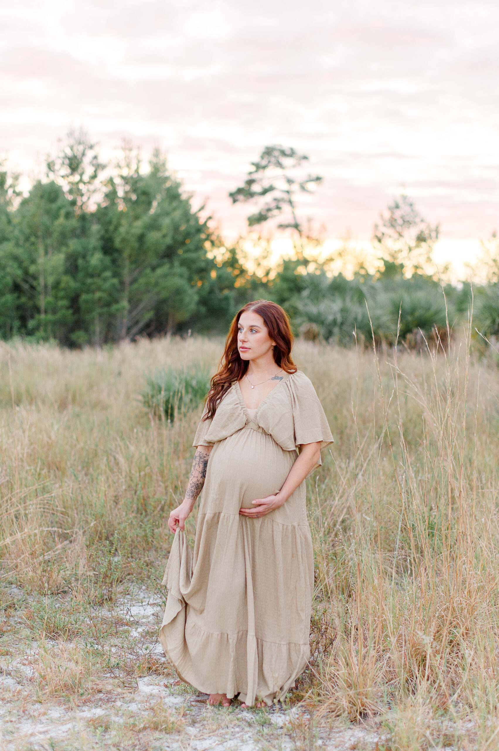 New mother looking over her shoulder while holding her belly and dress during her Orlando maternity photo session in a tall grass field Orlando prenatal yoga