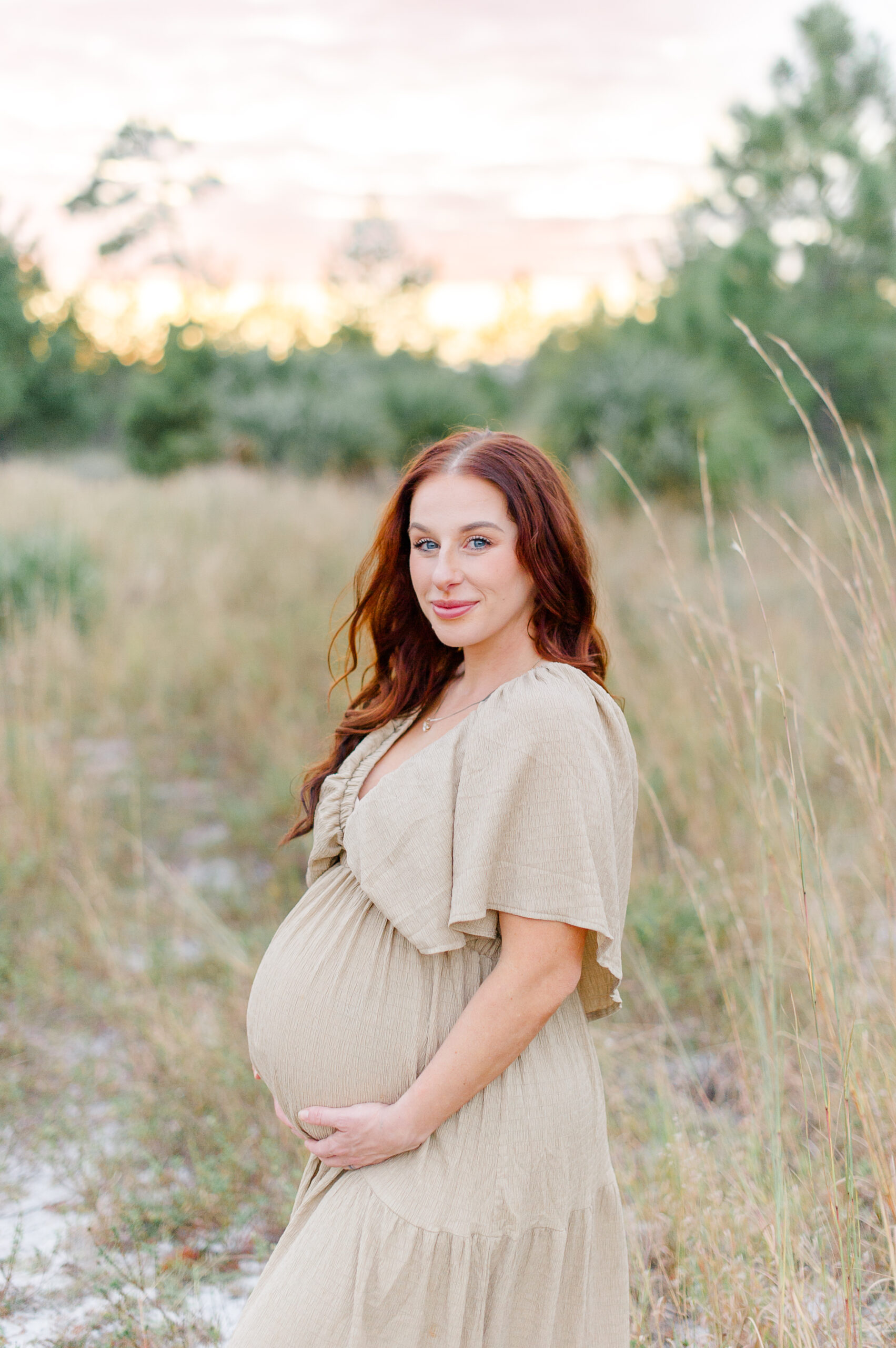 Expectant mother holding her belly while smiling softly at the camera during her photo session at golden hour