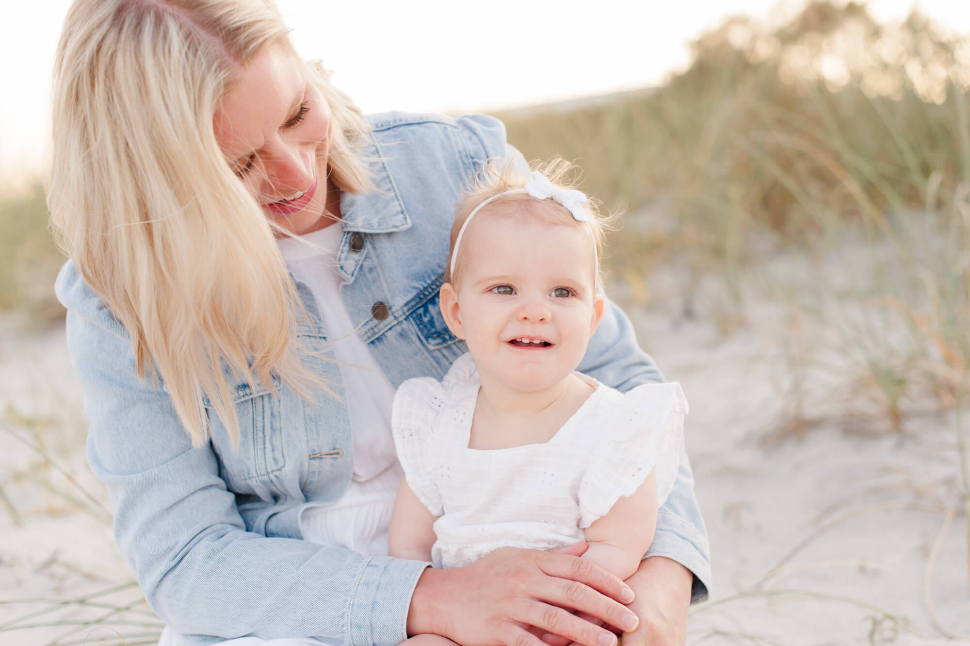 Mom holds daughter and smiles down at her while sitting in the dunes at sunset Viera Pediatric Dentistry