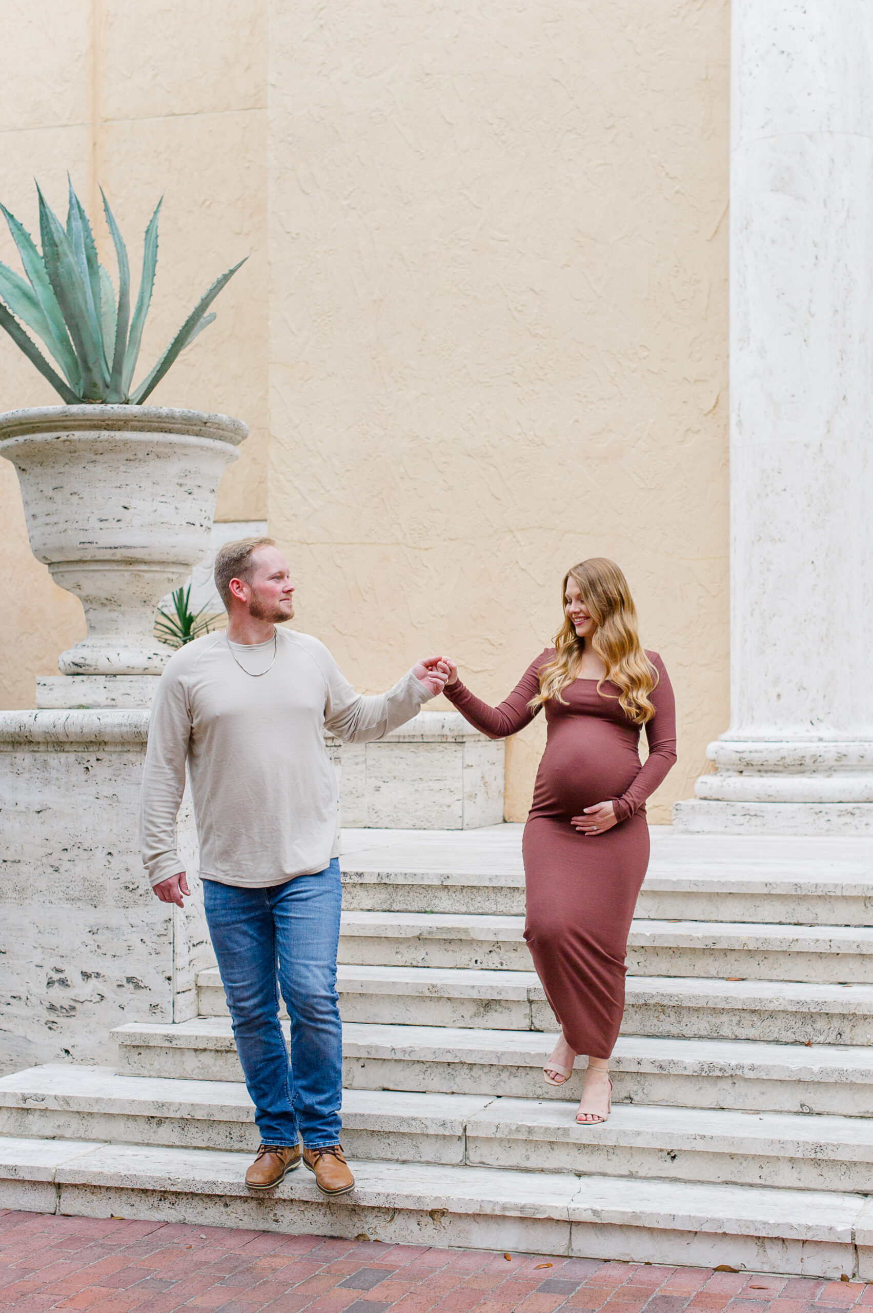 New father assists pregnant mother down steps while looking back at her lovingly during their maternity photo session.