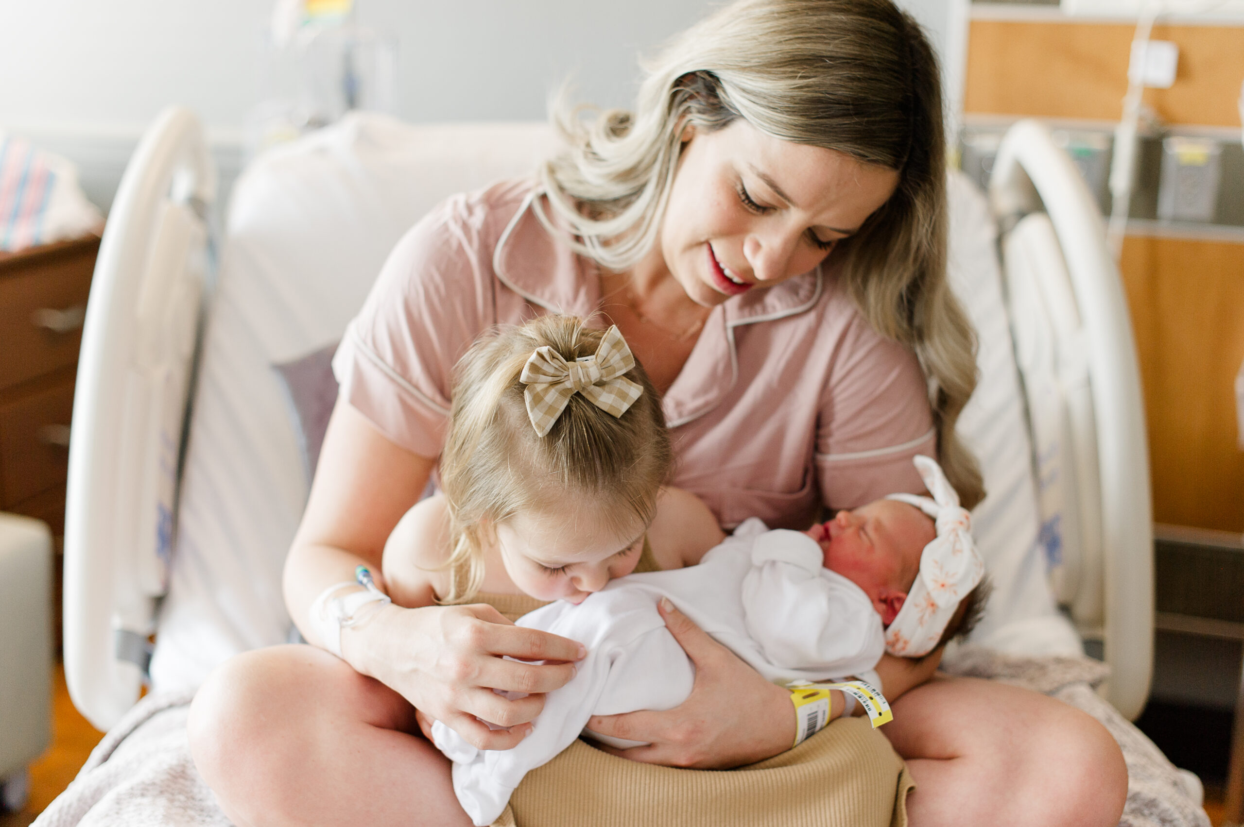Mother holds her sweet girls while her oldest kisses her newborn baby girl on the belly.